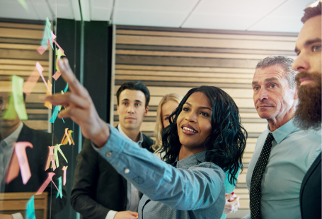 Attendees standing at team meeting looking at post-it notes on a glass wall.